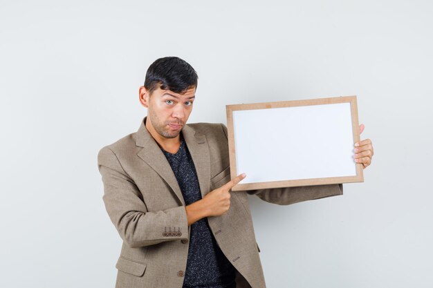 Young male pointing on white cardboard in grayish brown jacket and looking focused , front view.