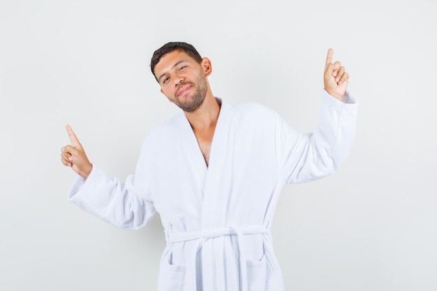 Young male posing while pointing up in white bathrobe and looking cheery. front view.