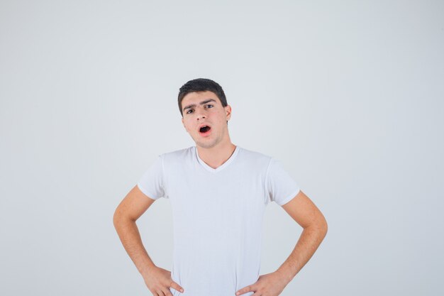 Young male posing with hands on waist while opening mouth in t-shirt and looking puzzled , front view.