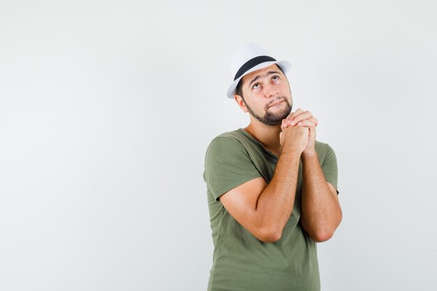 Young male praying with clasped hands in green t-shirt and hat and looking hopeful