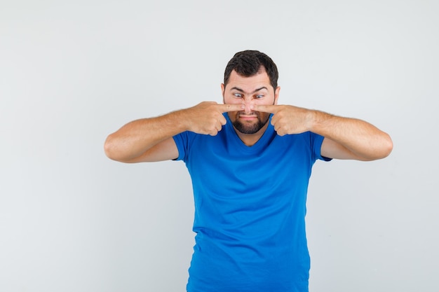 Young male pressing fingers on nose in blue t-shirt and looking weird