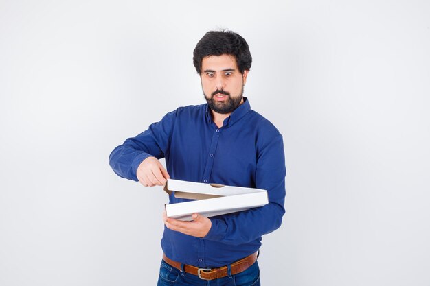 Young male pretending to open pizza box in shirt, jeans and looking amazed , front view.