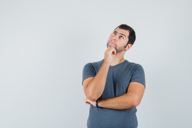 Young male propping chin on hand in grey t-shirt and looking hesitant  