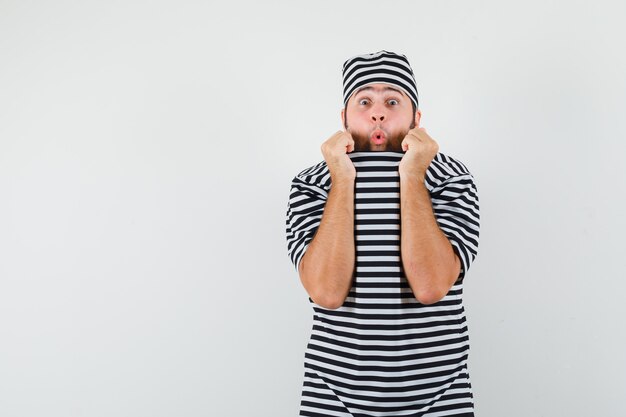 Young male pulling collar on face in t-shirt, hat and looking scared , front view.