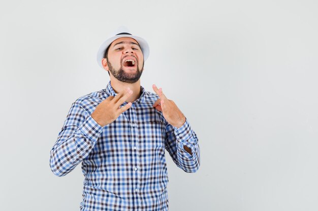 Young male pulling his shirt with fingers in checked shirt, hat and looking confident. front view.