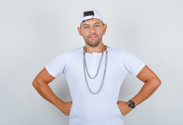 Young male putting fists on his waist in white t-shirt, cap, necklace and looking confident, front view.