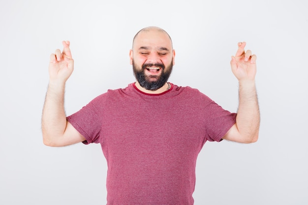 Free photo young male raising crossed fingers up while smiling in pink t-shirt and looking joyful. front view.