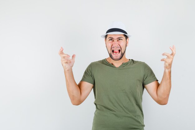 Young male raising hands in aggressive manner in green t-shirt and hat and looking furious