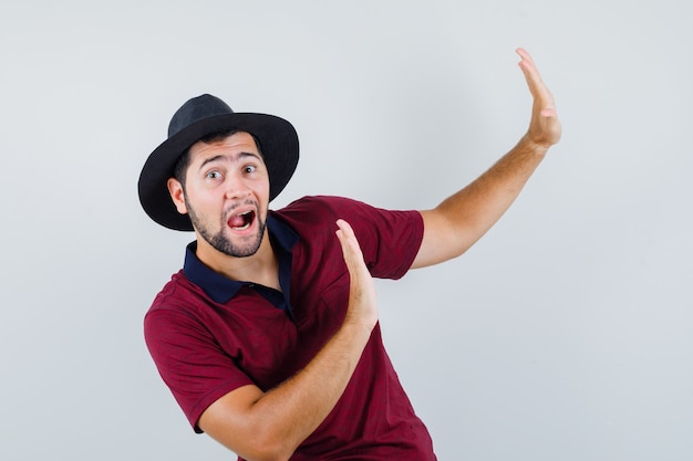 Young male raising hands to defend himself in t-shirt, hat and looking scared , front view.