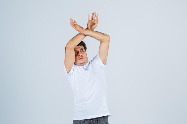 Young male raising hands to defend himself in white t-shirt, pants and looking scared , front view.