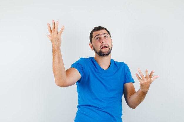 Young male raising hands in helpless manner in blue t-shirt and looking perplexed