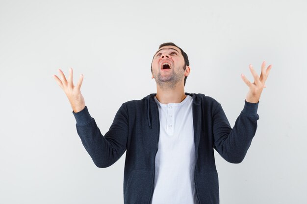 Young male raising hands while looking up in t-shirt, jacket and looking grateful. front view.