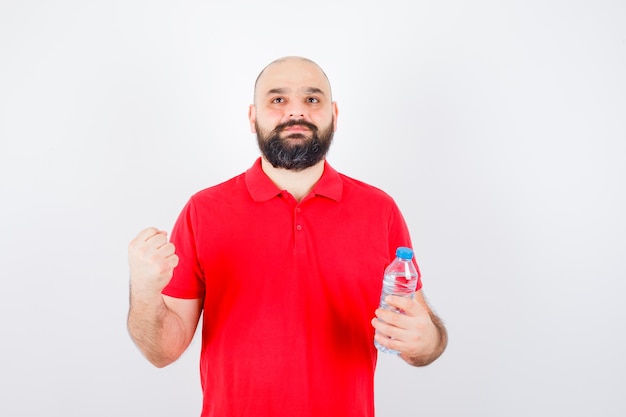 Young male in red shirt holding bottle while showing success gesture , front view.