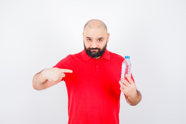 Молодой мужчина в красной рубашке, указывая на бутылку с водой и уверенно глядя, вид спереди.