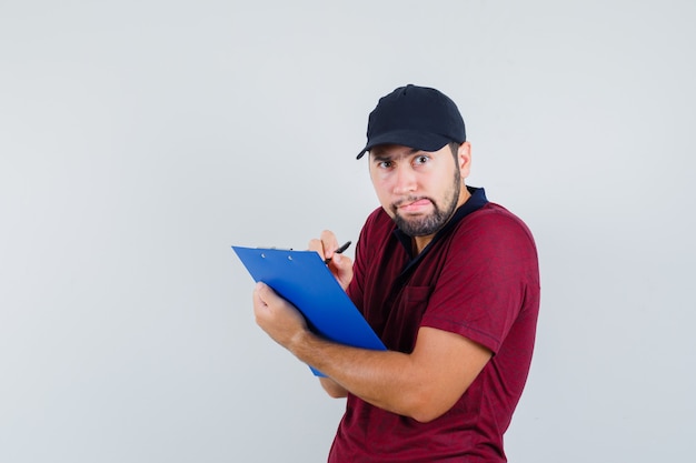 Young male in red t-shirt,black cap looking forward while writing something on his notebook and looking watchful , front view.
