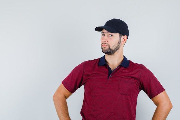 Young male in red t-shirt,black cap standing and looking weird , front view.