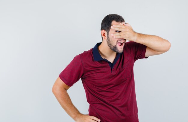 Young male in red t-shirt covering his face with hand while looking through fingers and looking funny , front view.