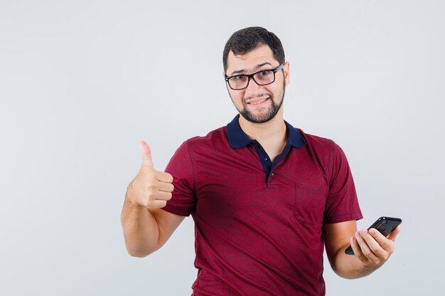 Young male in red t-shirt,glasses showing thumb up while holding phone and looking glad , front view.