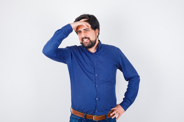 Young male in royal blue shirt holding hand on his head while smiling and looking merry , front view.