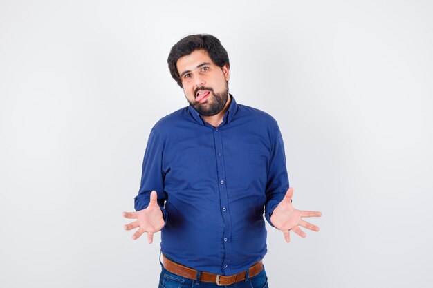 Young male in royal blue shirt opening his palms while sticking tongue out and looking weird , front view.