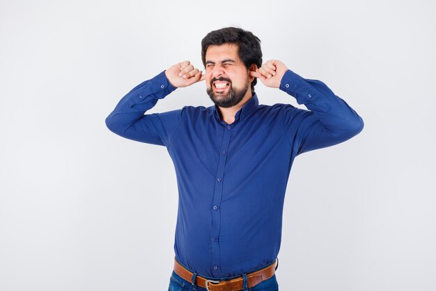 Young male in royal blue shirt plugging ears with fingers and looking stressful , front view.