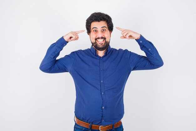 Young male in royal blue shirt pointing at his head while smiling and looking cheery , front view.