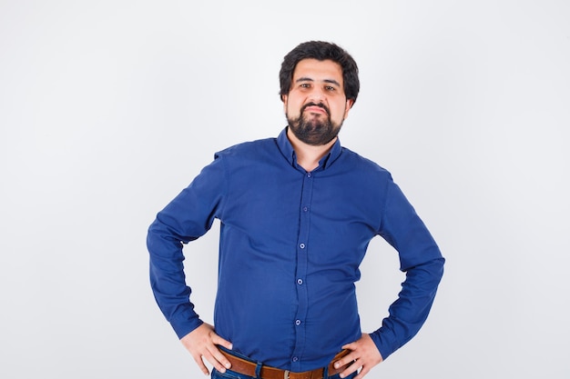 Young male in royal blue shirt standing with hands on waist and looking displeased , front view.