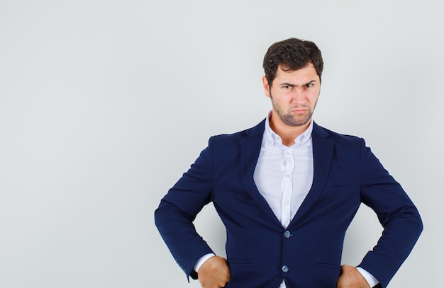 Young male scowling with hands on waist in suit and looking gloomy , front view.