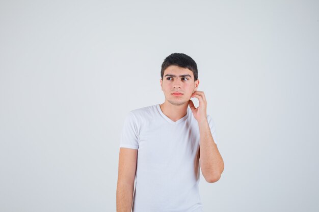 Young male scratching head while looking aside in t-shirt and looking pensive , front view.