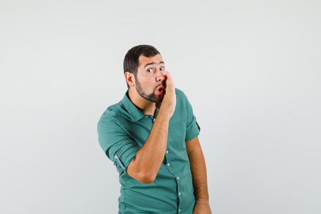 Young male secretly talking behind his hand in green shirt and looking omniscient , front view.