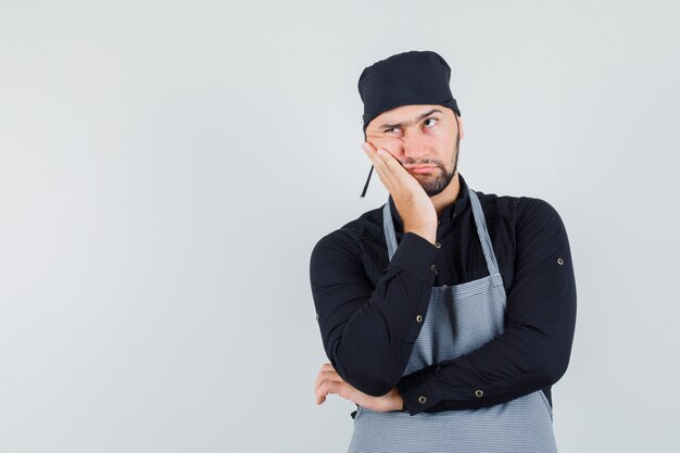 Young male in shirt, apron leaning cheek on raised palm and looking thoughtful , front view.