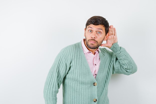 Young male in shirt, cardigan with hand behind ear and looking focused , front view.