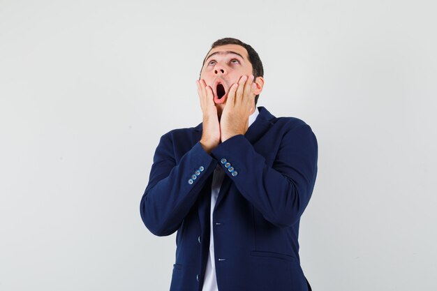 Young male in shirt and jacket holding hands on cheeks and looking surprised