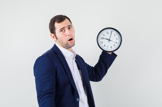 Young male in shirt and jacket holding wall clock and looking surprised