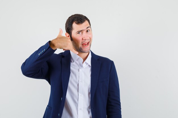 Young male in shirt and jacket showing phone gesture and looking cool