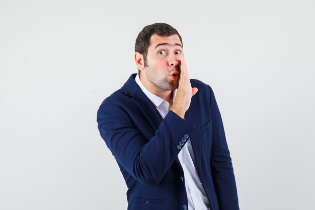 Young male in shirt and jacket telling secret behind hand and looking careful