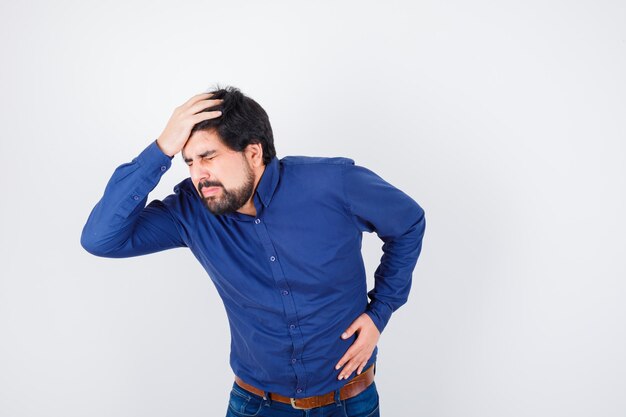 Young male in shirt, jeans holding hand on head and looking unwell , front view.