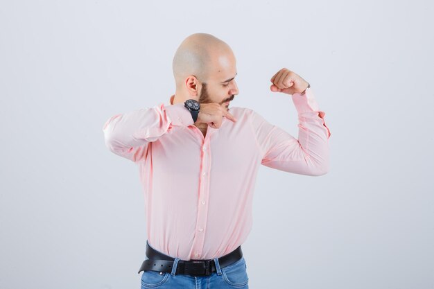 Young male in shirt, jeans pointing muscles of arm and looking proud , front view.