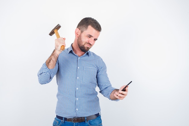 Молодой мужчина в рубашке, джинсах делает вид, что бьет молотком по мобильному телефону и выглядит серьезным, вид спереди.
