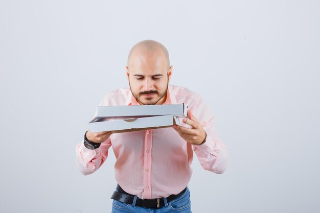 Young male in shirt, jeans smelling opened pizza box and looking delighted , front view.