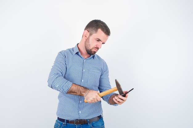 Young male in shirt, jeans striking mobile phone with a hammer and looking serious , front view.