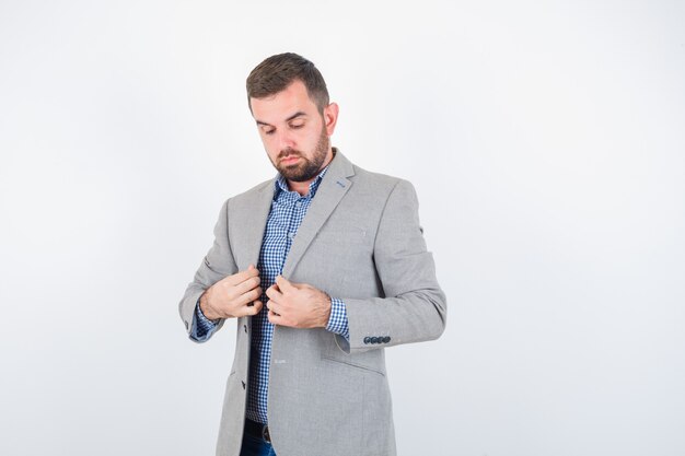 Young male in shirt, jeans, suit jacket holding lapels while posing and looking serious , front view.