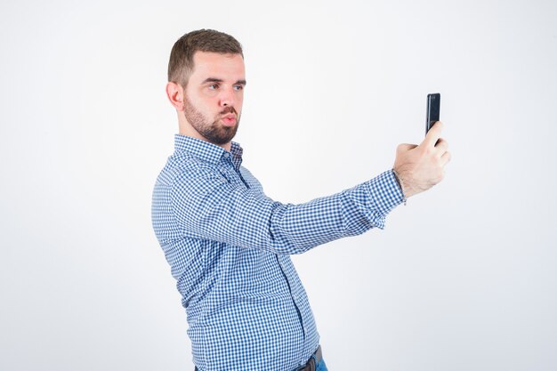 Young male in shirt, jeans taking a selfie while pouting lips and looking cute , front view.