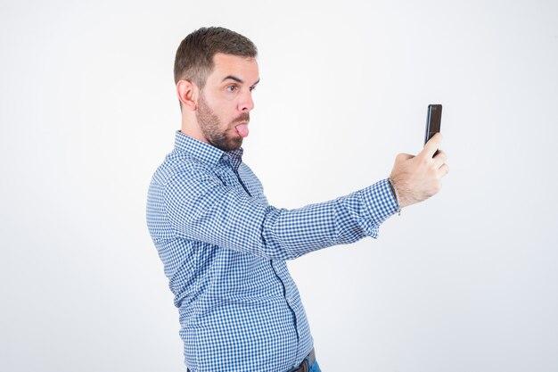 Young male in shirt, jeans taking a selfie while sticking out tongue and looking funny , front view.