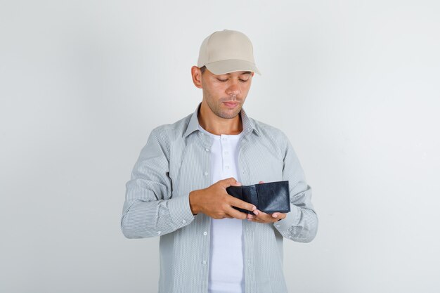 Молодой мужчина в рубашке с кепкой смотрит в открытый кошелек и смотрит осторожно