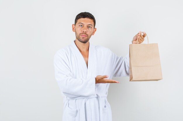 Young male showing craft paper bag in white bathrobe and looking strict. front view.