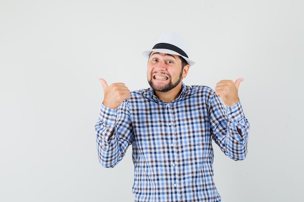 Молодой мужчина показывает двойные пальцы вверх в клетчатой рубашке, шляпе и выглядит веселым, вид спереди.