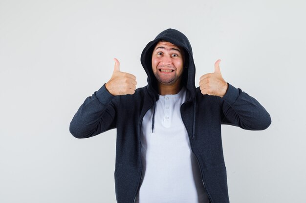 Молодой мужчина показывает двойные пальцы вверх в футболке, куртке и выглядит веселым, вид спереди.