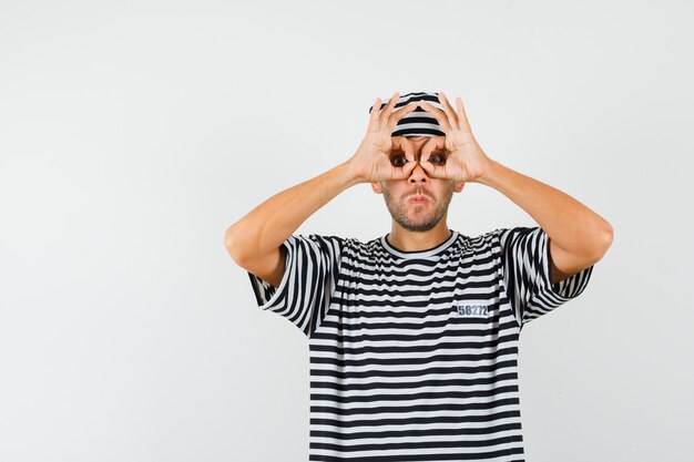 Young male showing glasses gesture in striped t-shirt hat and looking focused 