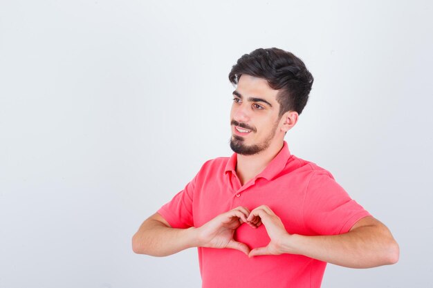 Young male showing heart gesture in pink t-shirt and looking confident. front view.